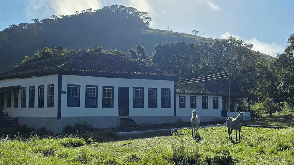 Fachada do casarão colonial da Fazenda Santa Clara com dois cavalos no gramado e o morro ao fundo