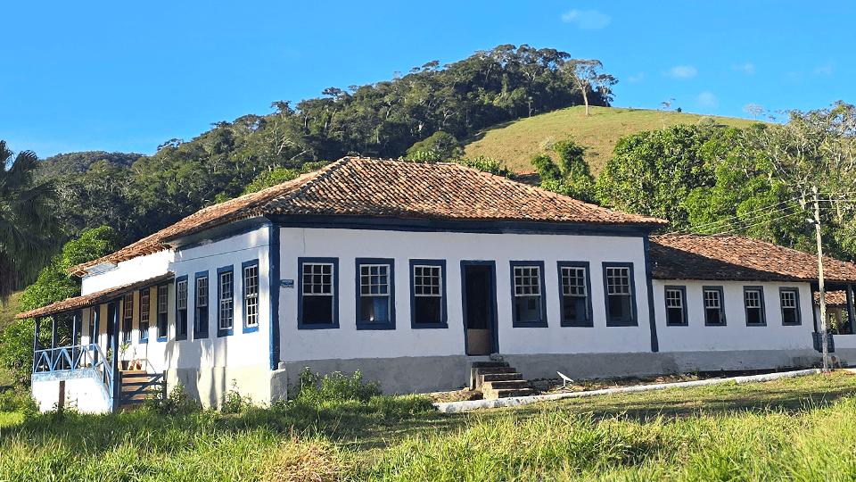 Casarão colonial branco da Fazenda Santa Clara sob céu azul, com o morro verde atrás